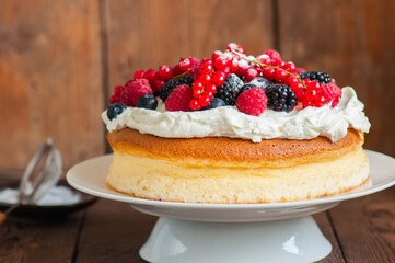 Close up of homemade fluffy Japanese cotton cheesecake decorated with fresh forest berries on a white cake stand on  a wooden background. Copy space.
