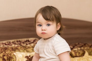 portrait of adorable baby girl with big blue eyes and long eyelashes, indoors. child sitting on the bed and looking into the camera. happy family concept