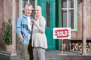happy senior couple holding keys of their new house and standing on porch