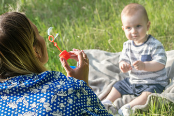 Young mather inflating soap bubbles for Surprised baby boy sitting on grass