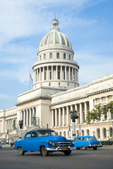 Fototapeta na wymiar Brightly colored classic American cars serving as taxis pass on the main street in front of the Capitolio building in Central Havana, Cuba