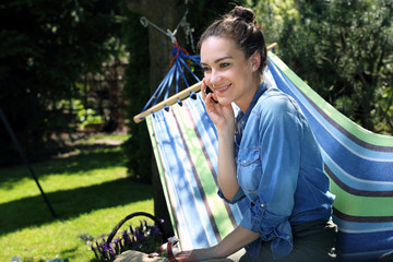 Wakacyjny relaks. Młoda atrakcyjna kobieta wypoczywa na hamaku w ogrodzie rozmawiając przez telefon