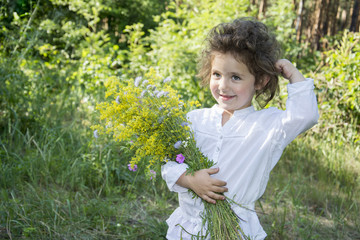 In summer, in the forest, a small curly girl holds a flower and laughs.