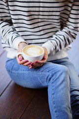 Woman with cup of cappuccino in hands sitting on the wooden floor