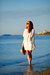 Beautiful woman in white clothes with orange juice, sunglasses and hat in hand on the beach