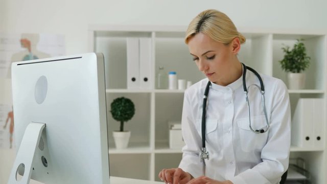 Young caucasian female doctor with stethoscope sitting at the table in office. Woman doctor typing on the computer keyboard, looking to the camera and smiling. Indoor.
