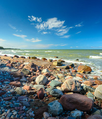 Fototapeta na wymiar Sunny day with blue sky on the beach with many stones. Waves on the background.