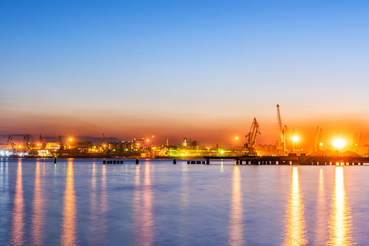 Type of night port. Working freight cranes, a ship at the pier, work in the port. Night photo view of the loading. Beautiful reflection of lights in the sea water.
