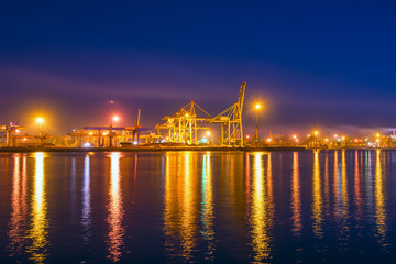 Type of night port. Working freight cranes, a ship at the pier, work in the port. Night photo view of the loading. Beautiful reflection of lights in the sea water.
