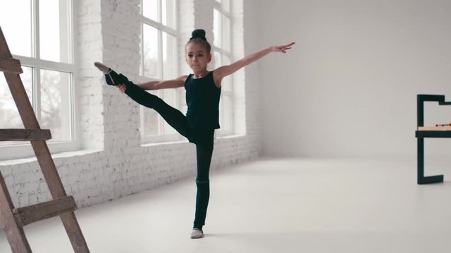 Cute little girl with twisted hair in a black sport uniform does the split in a ballet studio. Training time, keeping balance. Cheerful mood. Active lifestyle. White walls, modern studio interior.