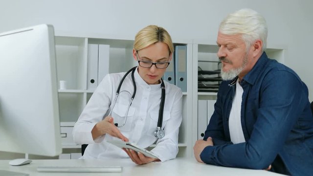 Caucasian female serious doctor explaining something to midle aged male patient using tablet sitting at desk in office. Indoor.