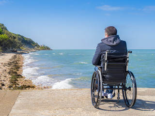 Disabled young man sitting in a wheelchair and looks at the sea.