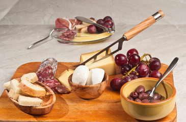  Gruyere cheese, grapes and salami and a bowl of black olives, a wood bowl of mozzarella cheese, a wood bowl of rustic italian bread,  glass of red wine, cheese knife on a wood serving board