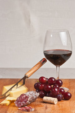 Gruyere cheese, grapes and salami with a glass of red wine , the cork and a cheese knife on a wood serving board