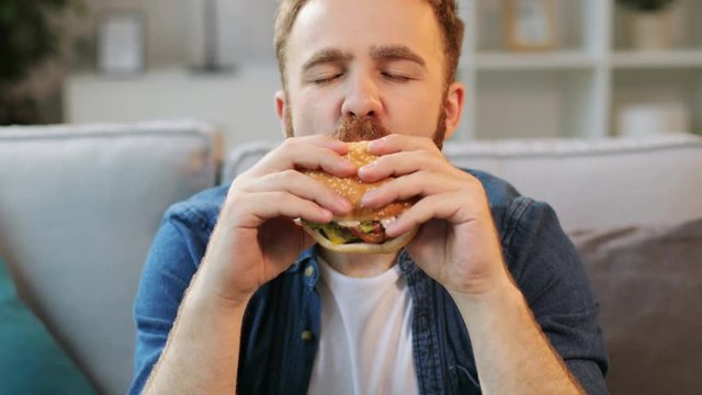 Portrait of young man eating junk food humburger in front of camera on living room background. Close up.