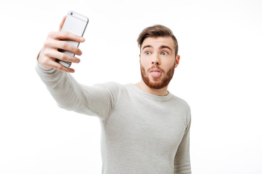Surprised casual young man taking selfie and showing tongue over white background