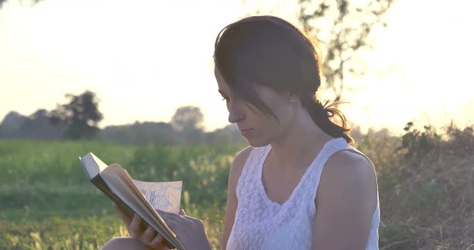 On a sunny day, a beautiful young student girl reads a book in nature (in a park, on a field) against a background of grass and sky. Concept ecology, clean air, summer, spring, grass, study, education