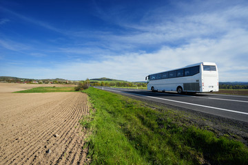 The white bus traveling on the road around a sown field in a rural landscape with a village in the background