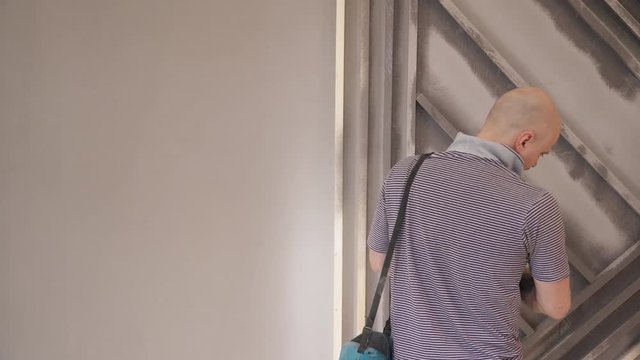 Master makes repairs and uses spray gun to wash metal shelves.Young European man in gray T-shirt with tool hanging from his shoulder washes crossbeams on the wall. He skillfully conducts the device