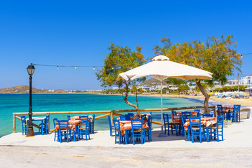 Tables with chairs in typical Greek tavern with view on sea bay. Naxos Island. Greece. - obrazy, fototapety, plakaty