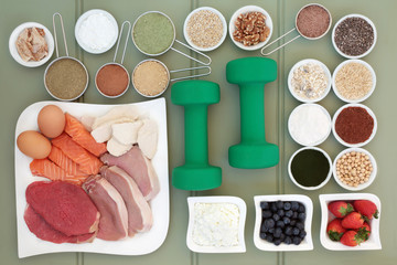 Super food for body builders with dumbbells, meat, fish, fruit, dairy, dietary supplement powders, grains, cereals, pulses, seed and herbs on green wood background.