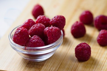 Fresh and slightly imperfect raspberries in and spilled around small glass bowl on wooden cutting board