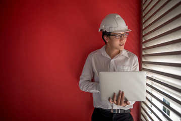 Young Asian Engineer or Architect dressed in white shirt and protective helmet working with laptop computer with red wall in background. Engineering, Architecture and building construction concepts