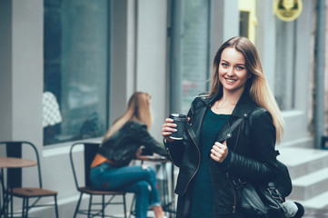 Fototapeta na wymiar Take away coffee. Beautiful young urban woman wearing in black stylish clothes holding coffee cup and smiling while walking along the street. Student's coffee break after study. Fashion lifestyle.