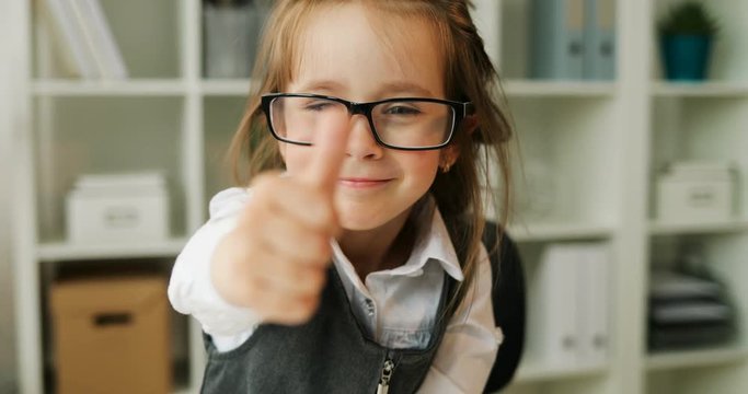 Beautiful young firl in formalwear with glasses looking at camera and showing thumb up on office background. Indoor. Close up.