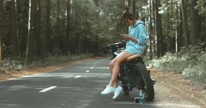 CINEMAGRAPH - seamless loop. Young Caucasian girl with brown hair sits on a motorbike on a forest road, checking her phone. 4K UHD RAW edited footage