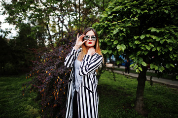 Fashionable woman look with black and white striped suit jacket, leather pants and sunglasses posing against bushes at street. Concept of fashion girl.