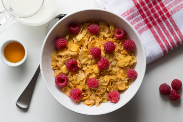 Breakfast cereal of cornflakes and raspberries, honey and milk on white table