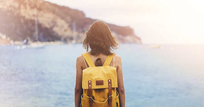 Back view hipster girl with backpack in sand coastline on nature landscape, mock up. Traveler on background beach seascape and horizon mountain. Tourist look on blue sun ocean, summer relax lifestyle