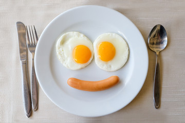 Smiling face frying eggs breakfast on a white plate background with spoon,knife and fork on a fabric table, breakfast serve for kid.