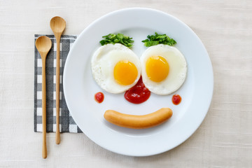Top view of fried egg smile face on white plate with wooden spoon on white fabric table, breakfast serve for kid.