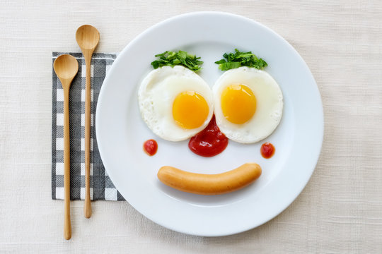 Top View Of Fried Egg Smile Face On White Plate With Wooden Spoon On White Fabric Table, Breakfast Serve For Kid.