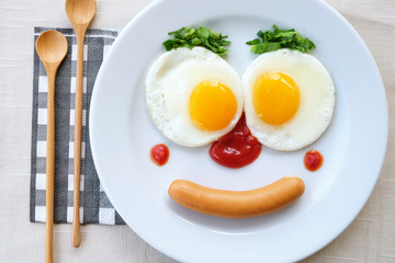 Top view of fried egg smile face on white plate with spoon knife and fork on purple fabric table, breakfast serve for kid.