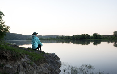 Female traveller sits on the stone above the river. Sunset time. Hills and forest on the banks