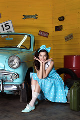 Young beautiful woman in blue dress with polka dots with bow on head smiling looking at camera and holding hands under head near blue car