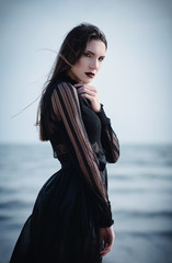 Beautiful sad goth girl in black dress standing on sea beach