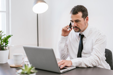 Middle aged man making call and using his laptop while working from home office