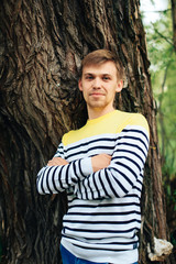 Portrait of a young guy with a beard against the background of the bark of an old tree in a yellow striped swish, summer nature