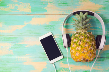 Pineapple with headphones and smart phone on wooden background. Tropical summer vacation and beach party concept. View from above