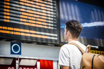 Young man traveling, reading big electronic train timetable in railway station, seen from behind
