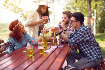 Young people play cards and toast with beer at table in nature