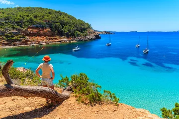 Foto op Aluminium Young woman tourist standing on rock cliff edge and looking at beautiful Cala Salada bay famous for its azure crystal clear sea water, Ibiza island, Spain © pkazmierczak