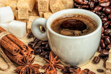 A white coffee cup on a wooden table, surrounded by coffee beans, sugar cubes, cinnamon sticks and anise flowers