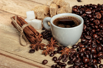 A white coffee cup on a wooden table, surrounded by coffee beans, sugar cubes, cinnamon sticks and anise flowers