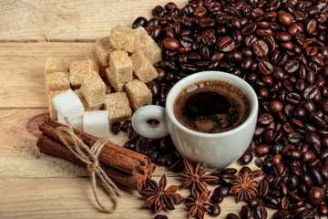 A white coffee cup on a wooden table, surrounded by coffee beans, sugar cubes, cinnamon sticks and anise flowers