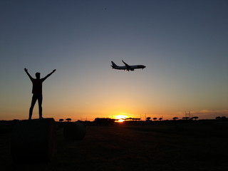 Fototapeta na wymiar Giovane ragazzo con le mani al cielo e aereo in atterraggio al tramonto